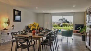 Un coin repas avec une table dressée pour quatre personnes, des chaises, un vase rempli de fleurs jaunes, une lampe sur pied et une grande fenêtre donnant sur un jardin.