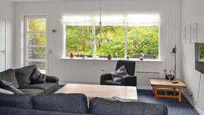 Un salon comprenant un canapé, un fauteuil et une table basse, de grandes fenêtres donnant sur un paysage verdoyant, et un téléviseur fixé au mur.