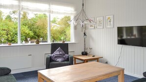 Un salon moderne avec une table basse en bois, un canapé gris et un fauteuil agrémenté d’un coussin violet. Un téléviseur est fixé au mur et une fenêtre offre une vue sur la verdure à l’extérieur.