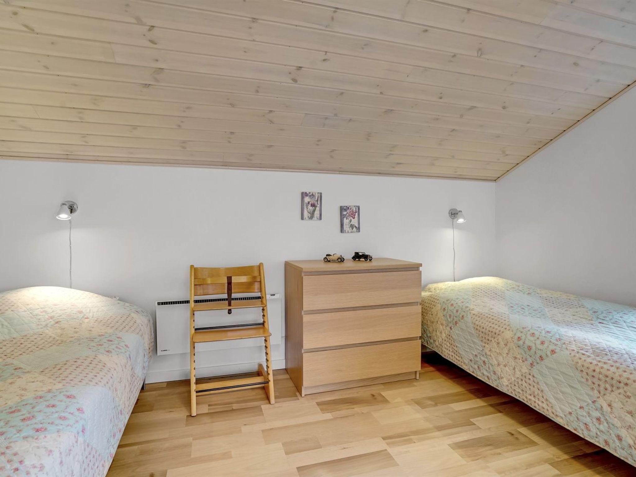 Un dormitorio con dos camas individuales, un aparador de madera, una silla y dos lámparas fijadas a la pared.