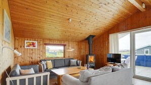 Interior acogedor de una cabaña con techo de madera, un sofá, una mesa de comedor, un televisor y una estufa de leña.