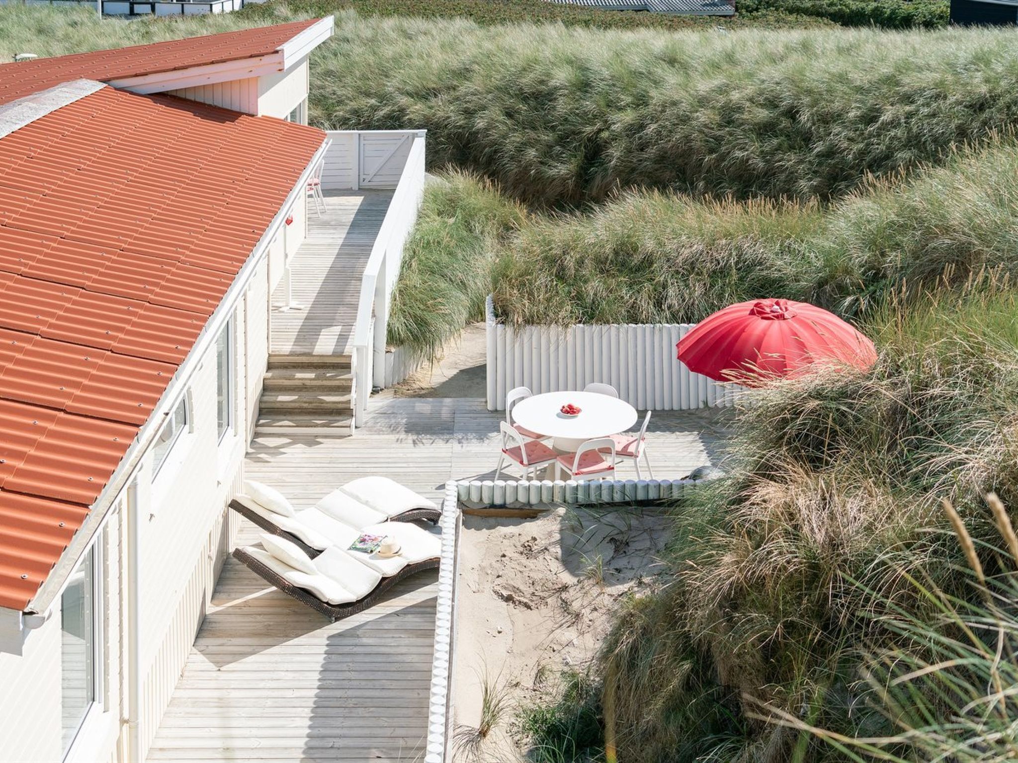 A beach house with a wooden deck, a red umbrella, and a white outdoor table and chairs.