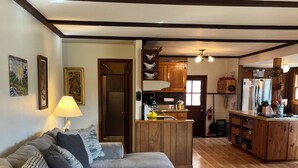 Ein Wohnzimmer mit Ecksofa, Hocker und einem runden Teppich. Es gibt eine Küchenzeile mit Holzschränken und einem Kühlschrank.