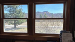 Ein Zimmer mit großem Fenster, das auf einen bewaldeten Bereich und einen Berg blickt. Am hölzernen Tisch stehen ein Korb und ein paar Papiere.