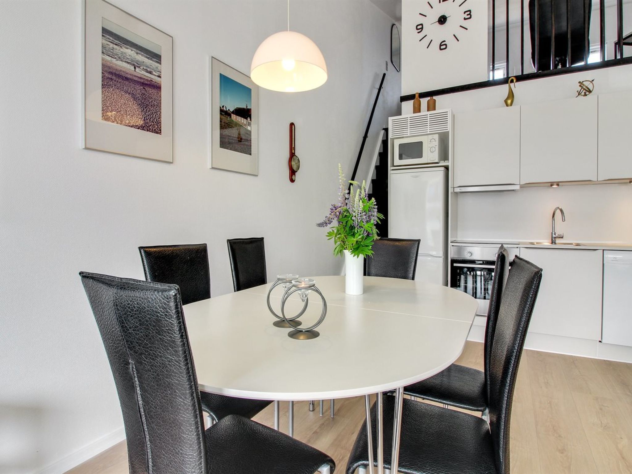 A modern dining area with a white table, black chairs, and a vase with flowers. There are framed pictures on the wall and a clock above the door.