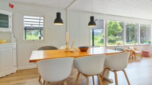 Un comedor amplio con una mesa de madera y sillas blancas, ventanas grandes con vistas a zonas verdes, y lámparas colgantes.
