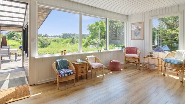 Una habitación iluminada por el sol con ventanas amplias, mobiliario de mimbre y vistas al verdor exterior.