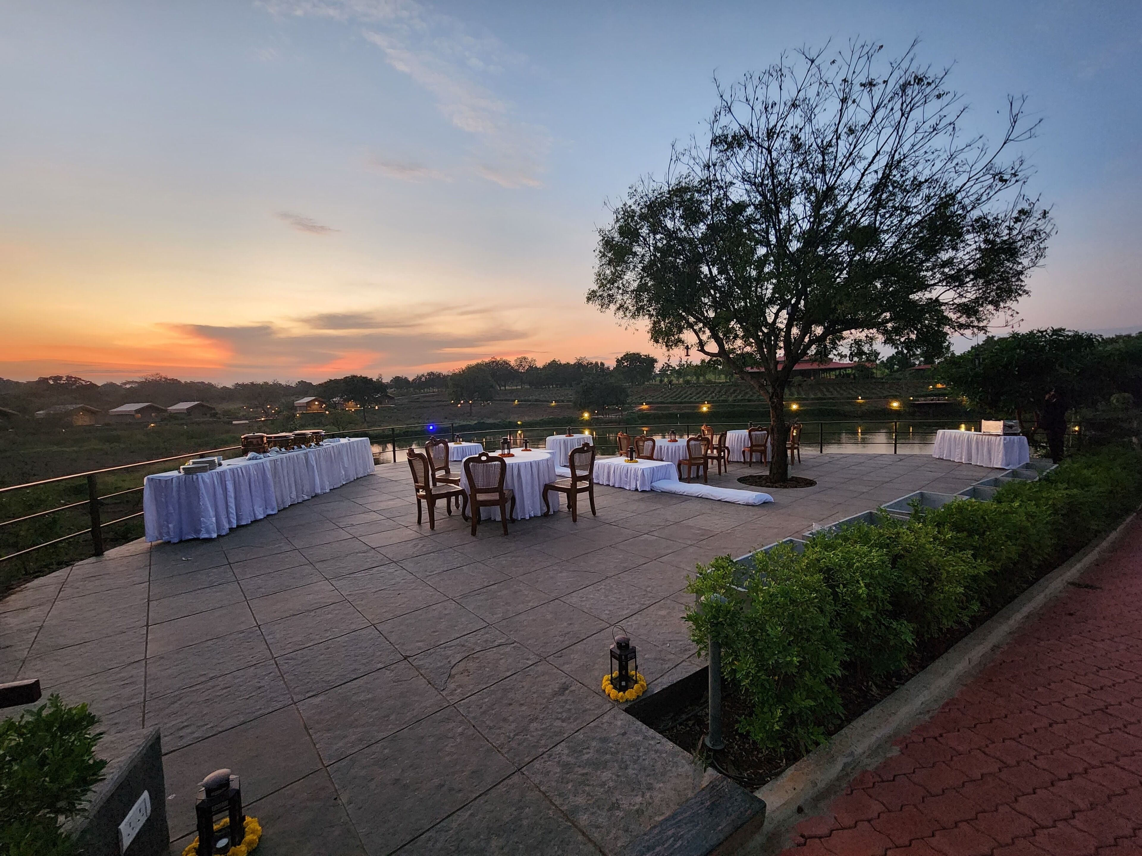 Un espace de restauration en plein air avec des tables rondes recouvertes de nappes blanches, entourées de chaises, le tout se dressant sur un décor pittoresque, avec un coucher de soleil se reflétant sur une étendue d'eau.