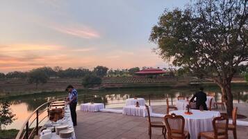 An outdoor dining area with tables set up for guests, overlooking a body of water and a distant building.
