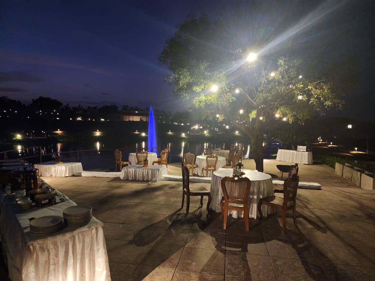 An outdoor dining area with round tables and chairs, set against a backdrop of a body of water and a blue-lit structure.
