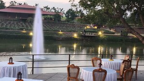 A serene outdoor dining area with tables and chairs by a calm water body, illuminated by ambient lighting. - The Hamlet (Kondapur)