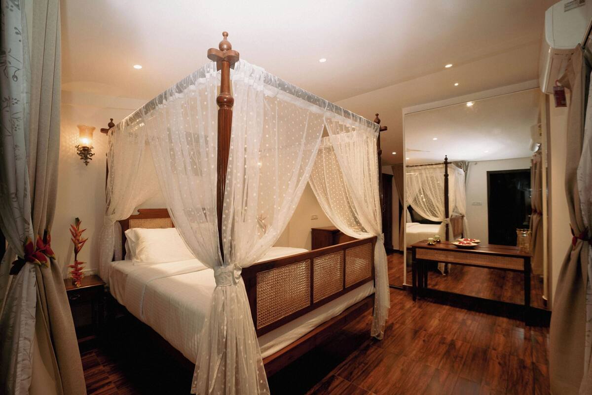 Una cama con dosel sobre cuatro columnas, con estructura de madera y ropa de cama blanca, una mesita de noche con lámpara y un espejo en la pared.