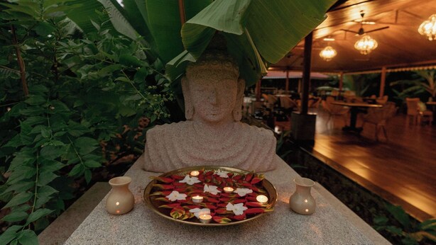 A Buddha statue with a plate of flowers and candles, surrounded by greenery and a wooden structure with hanging lights. - The Hamlet (Kondapur)