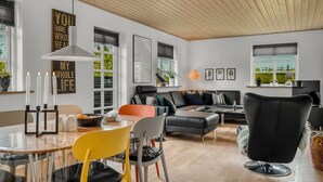 A modern living room with a wooden ceiling, a black leather sofa, a dining table with chairs, and a large window with blinds.