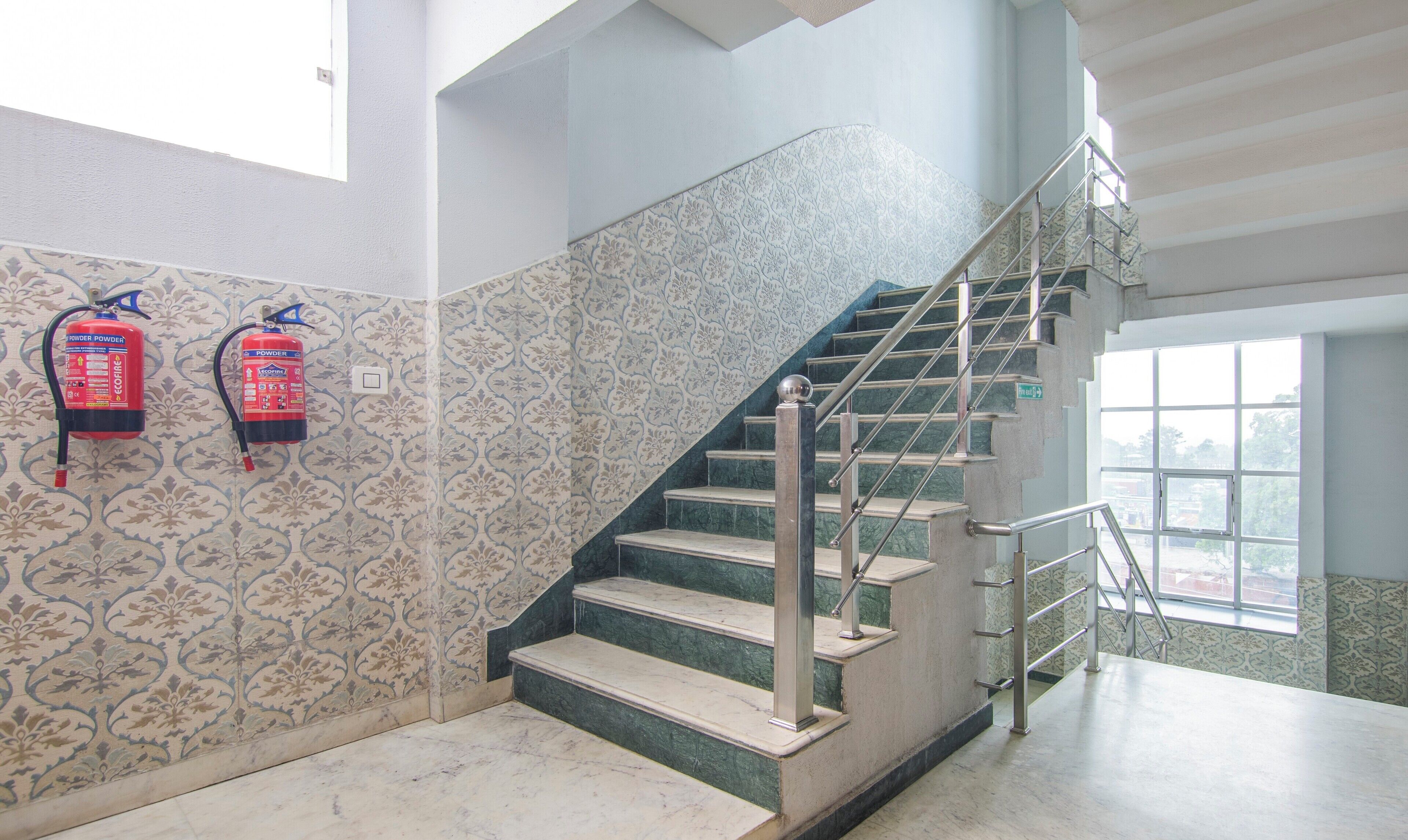 A staircase with marble steps and metal railings, fire extinguishers on the wall, and a window with a view of buildings.