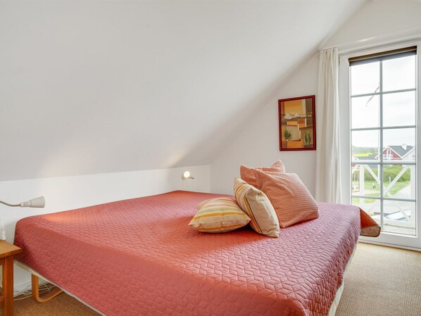 Une chambre à coucher avec une couvre-lit rouge matelassé, une table de chevet en bois, une lampe de chevet et une fenêtre donnant sur une maison et de la verdure.