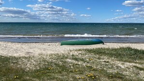 Une plage avec un kayak vert et une rive sablonneuse parsemée d’herbe clairsemée et de fleurs jaunes.