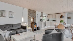 Una sala de estar moderna con un sofá, una mesa de centro y una chimenea. Se observa una zona de cocina con armarios y una mesa de comedor.