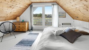 Un dormitorio con techo inclinado, una cama con almohadas, un aparador, una silla y vistas al exterior.