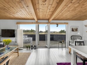 Un interior moderno con techo de madera, un televisor, una mesa de comedor y vistas a través de grandes puertas de vidrio.