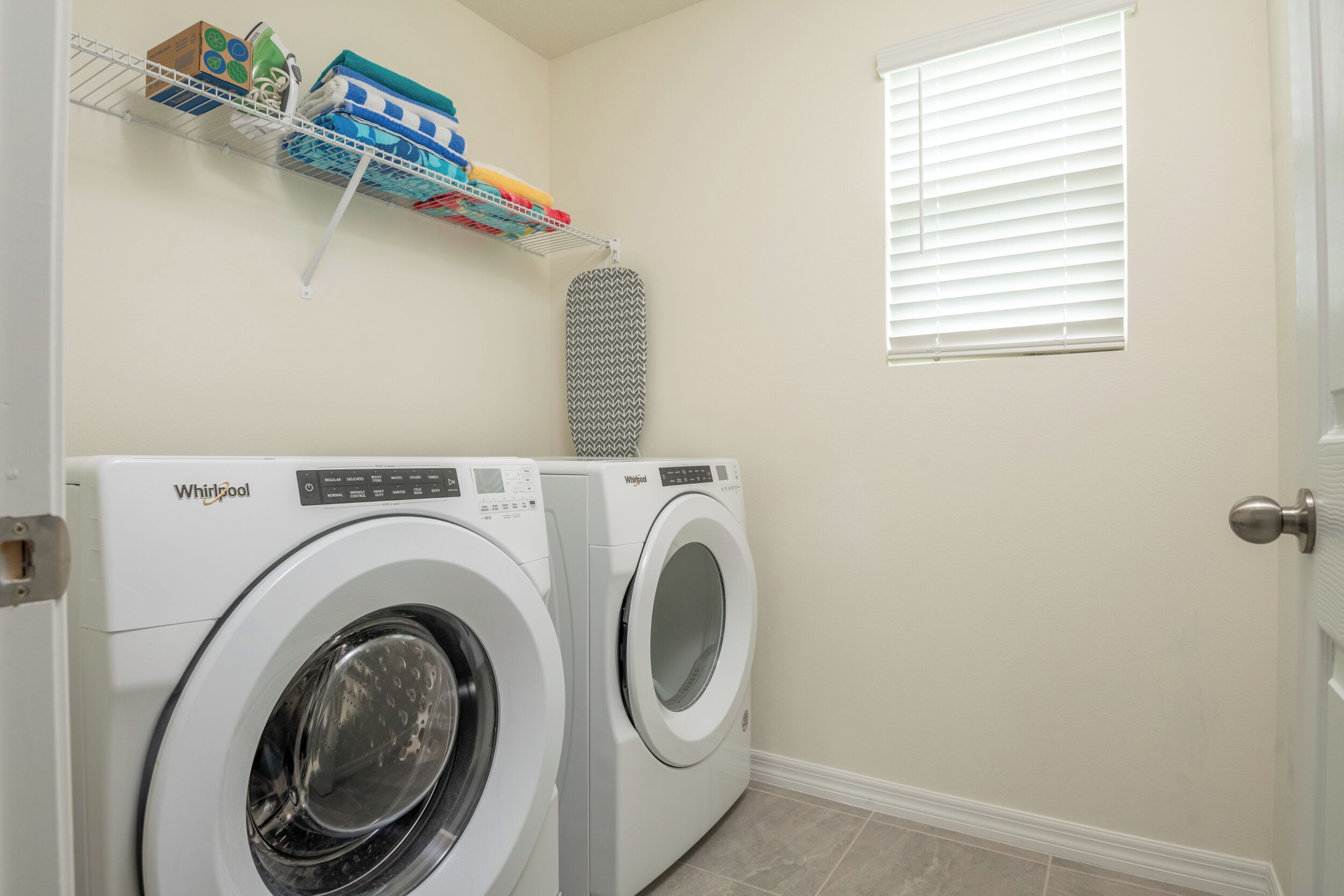 A laundry area with two Whirlpool washers, a shelf with cleaning supplies, and a window with blinds.