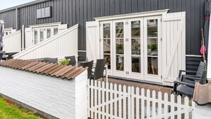 Ein modernes Gebäude mit weißen Außenwänden und einem dunkelgrauen Dach. Es besitzt einen weißen Zierzaun, eine Holzterrasse und eine Treppe, die zu einer weißen Tür führt.