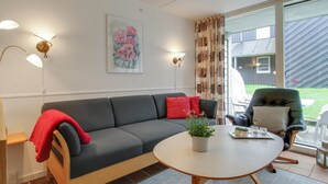 Een moderne woonkamer met een grijze bank, een ronde salontafel en een groene fauteuil. Er hangt een schilderij aan de muur en er is een raam met gordijnen.