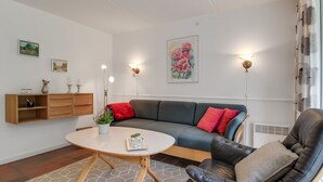 Een moderne woonkamer met een grijze bank, een zwarte lederen fauteuil, een ronde houten salontafel en een vloerkleed met een geometrisch patroon.