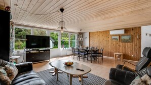 A spacious living room with a wooden ceiling, a large TV, a dining area with a table and chairs, and a black leather sofa.