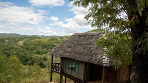 A thatched-roof hut with a balcony, surrounded by lush greenery and hills in the background.