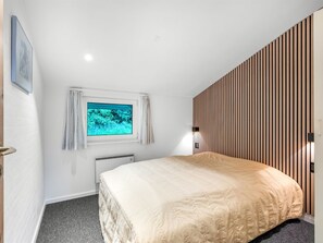 Une chambre à coucher avec un lit, une fenêtre avec des rideaux, un radiateur et un mur recouvert de lambris en bois.