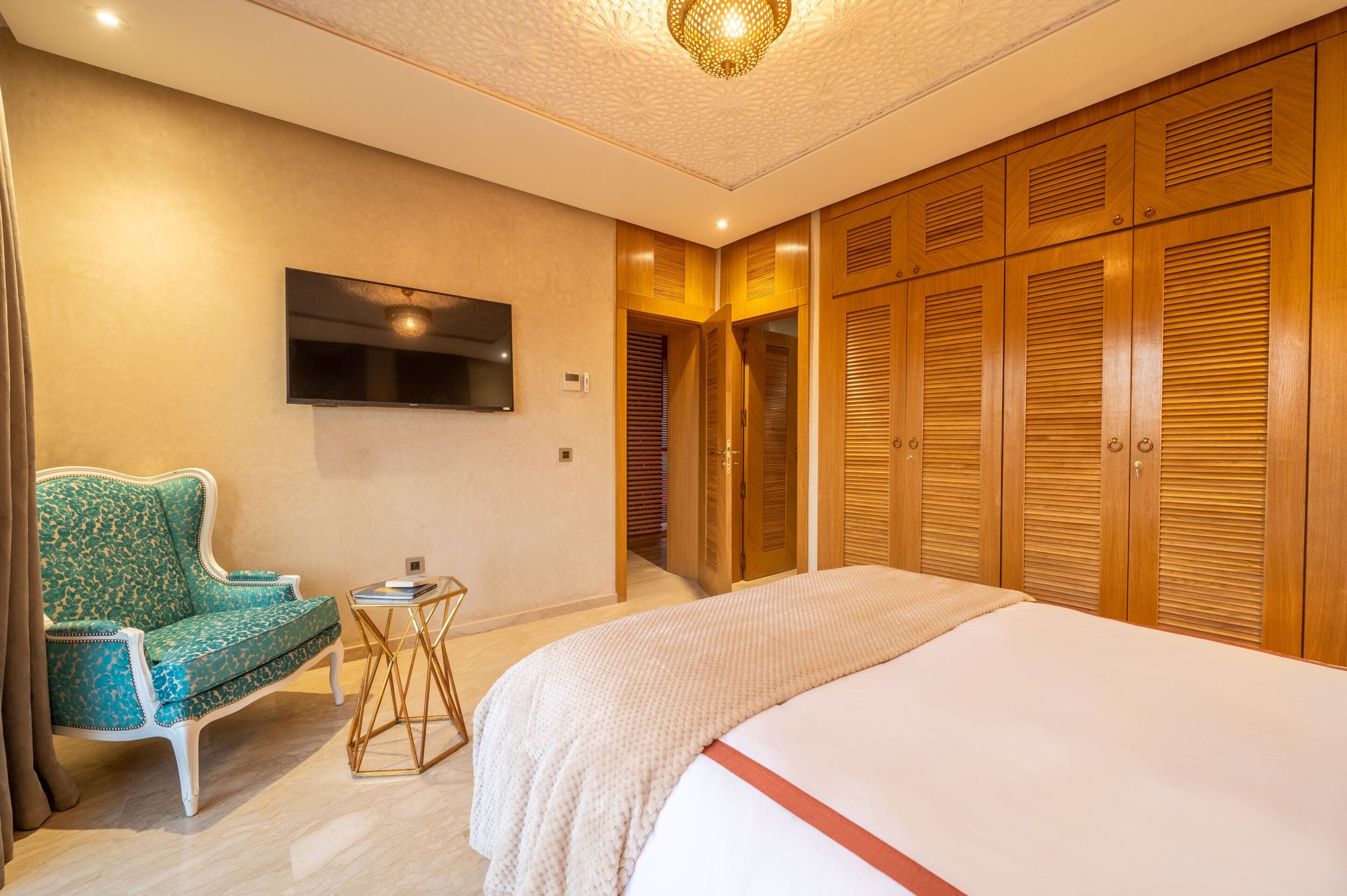 A hotel room with a bed, a TV mounted on the wall, a green patterned sofa, a small side table, and wooden closets.