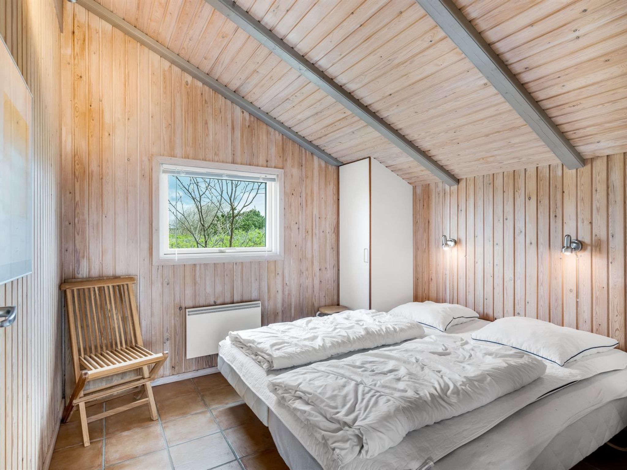 Une chambre avec des murs en bois, un lit, une chaise, une fenêtre et un radiateur.