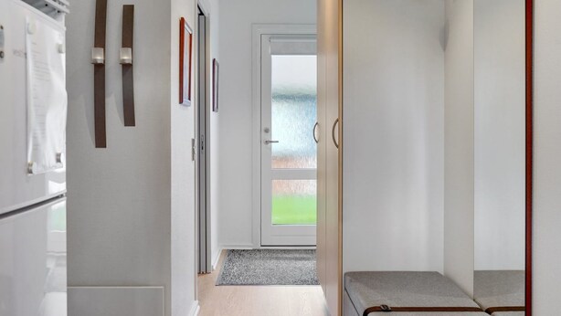 A narrow hallway with a door leading to a balcony, a small seating area, and a refrigerator on the left.