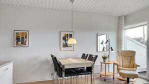 A modern dining area with a wooden table, black chairs, and a cream-colored armchair. There are two framed artworks on the walls and a pendant light above the table.