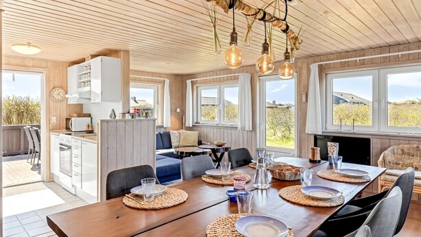 A modern kitchen with white cabinets, a dining area with a wooden table set for six, and large windows offering views of the outdoors.