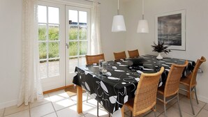 Ein Essbereich mit einem für sechs Personen gedeckten Tisch, umgeben von Korbstühlen, mit einer schwarzen Tischdecke mit weißen abstrakten Mustern. Zwei Pendelleuchten befinden sich über dem Tisch, und ein gerahmtes Bild hängt rechts an der Wand.