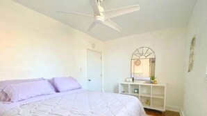 Un dormitorio con cama, ventilador de techo, puerta blanca y estantería blanca con varios objetos.