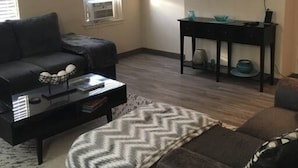 Una sala de estar con un sofá oscuro, una mesa de centro, un televisor en la pared y una mesita auxiliar.