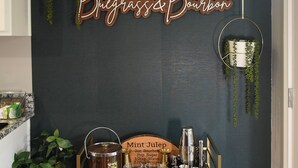 A bar area with a sign reading "Bleggrass & Bourbon," a wooden bar cart with glasses and bottles, and hanging plants.