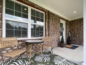 Un patio cubierto con dos sillas de mimbre y una mesa pequeña, un muro de ladrillo y un ventanal amplio.