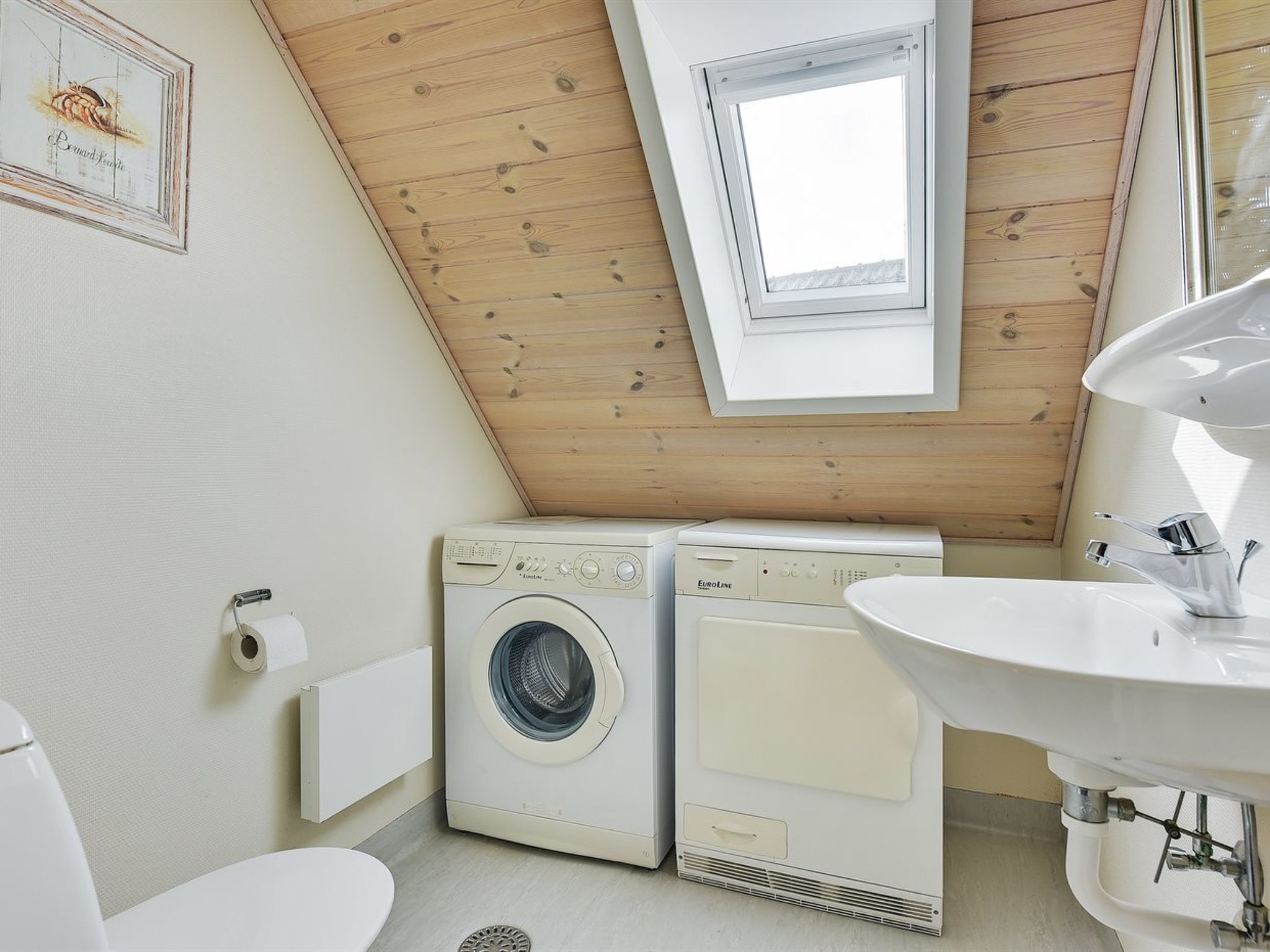 A compact bathroom with a sloped ceiling, a window, a washing machine, a sink, and a toilet.