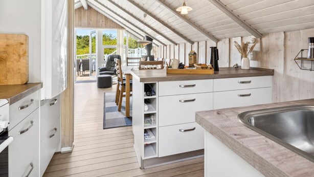Et moderne køkken med hvide skabe, en træbordplade og et stort vindue med udsigt til en terrasse.