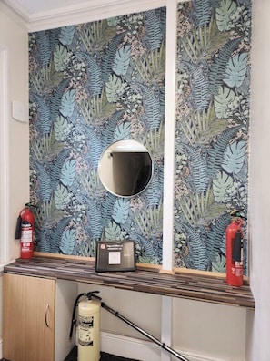 Features a unique floral wallpaper design, a round mirror, and a console table with a framed item and fire extinguisher.