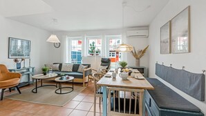 Una sala de estar moderna con un sofá, una mesa de comedor y sillas. Hay ventanas con vistas a edificios.