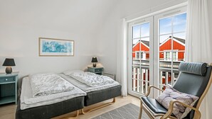 Un dormitorio con dos camas, una silla, una mesita de noche, una lámpara y una ventana con vista a edificios rojos.