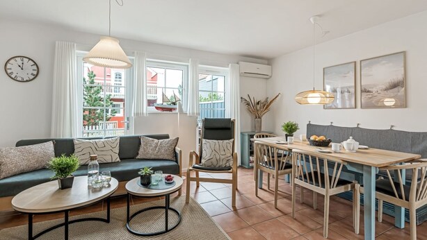 Una sala de estar moderna con sofá, mesa de centro y comedor. Hay un reloj en la pared y una ventana con vistas a edificios.