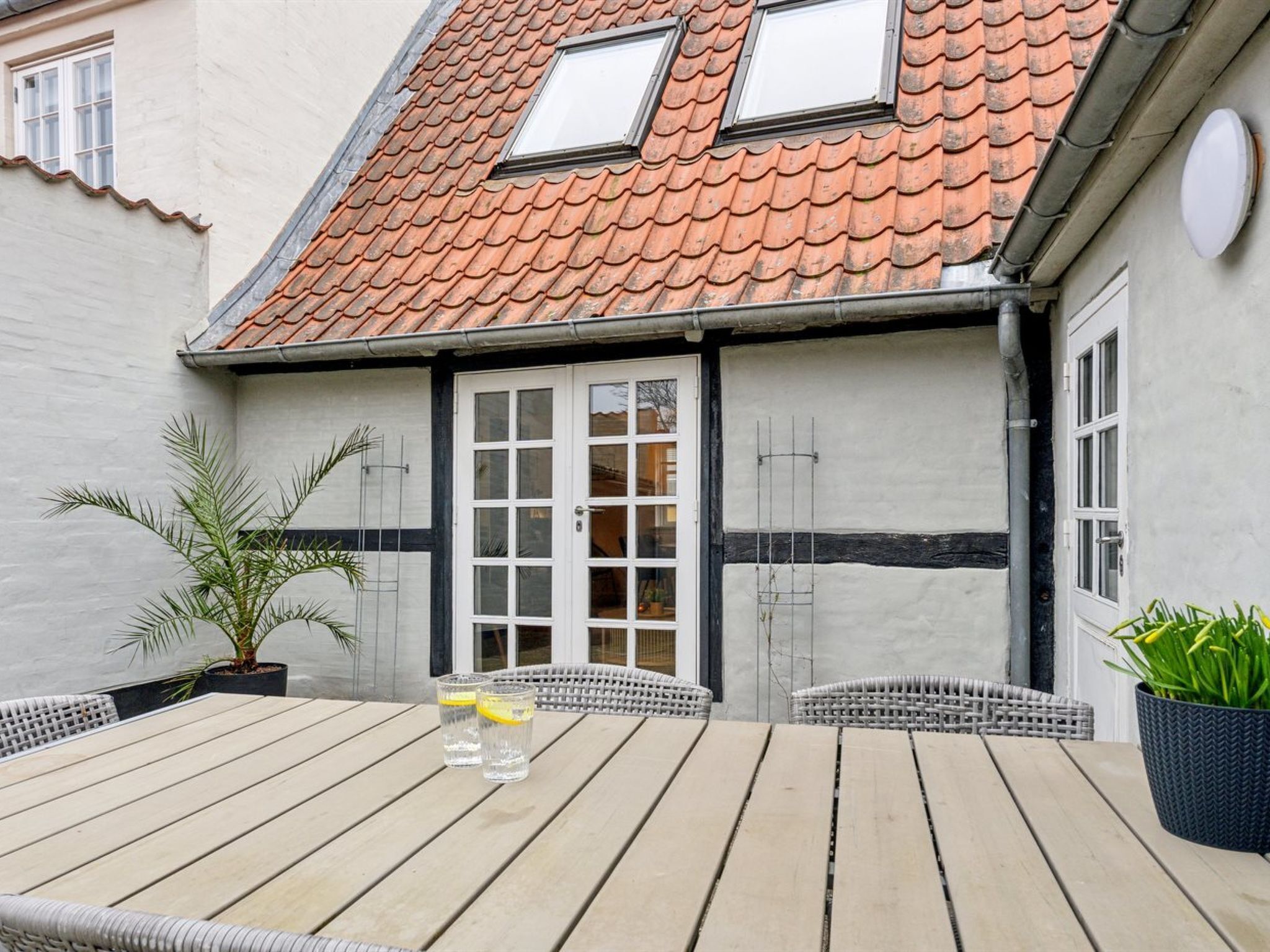 Une terrasse en bois avec une table et des chaises, un pot de fleurs et un bâtiment doté d’un toit en tuiles et de fenêtres.