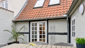 Een houten terras met een tafel, stoelen, een bloempot en een gebouw met een pannendak en ramen.