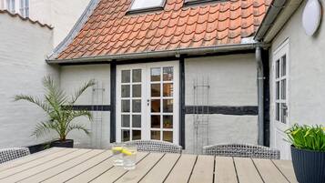 A wooden deck with a table and chairs, a potted plant, and a building with a tiled roof and windows.
